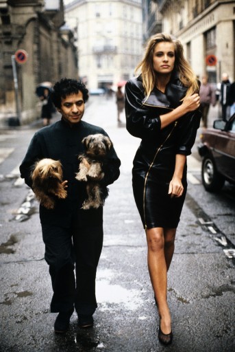 Fashion designer Azzedine Alaia holding his two Yorkshire terriers, Patapouf and Wabo, walking in Paris street with model Frederique who wears one of his creations, a black leather zippered dress, 1986. Courtesy of Arthur Elgort.